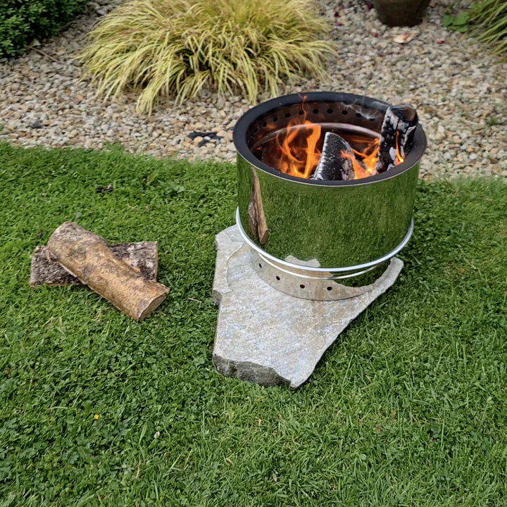 A portable stainless steel patio heater with a natural flame, placed on grass next to a kiln dried log.